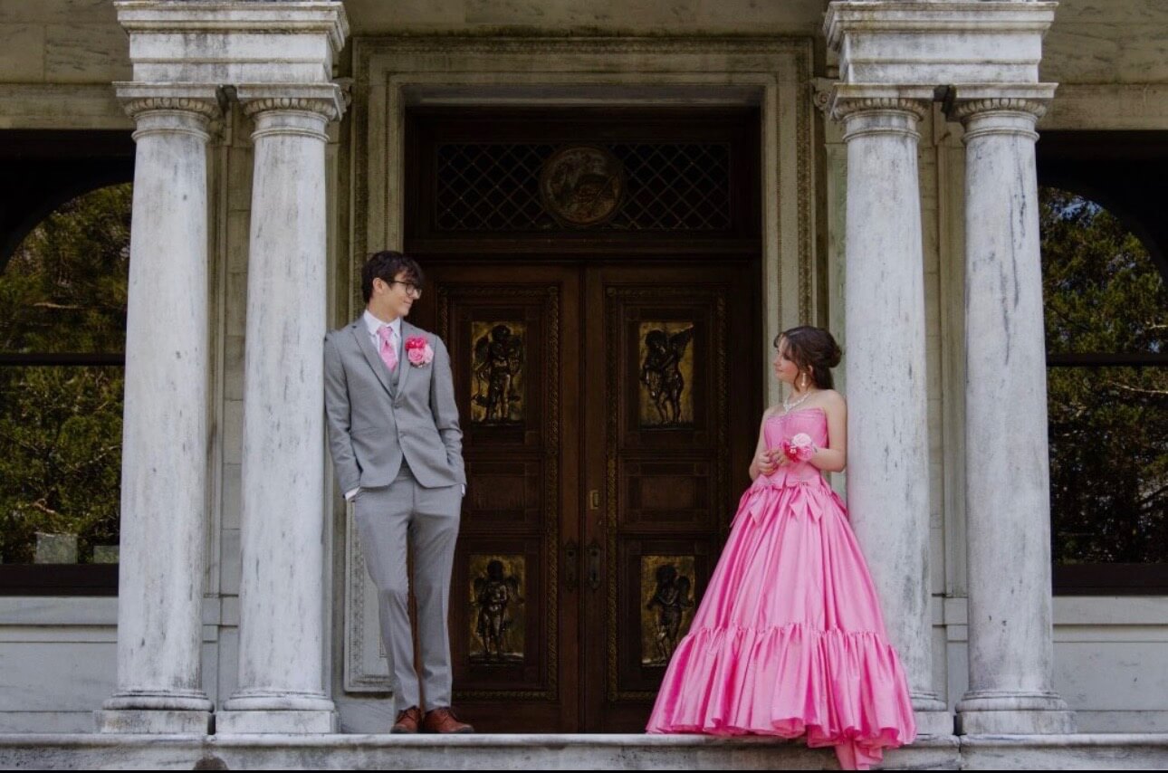 A couple poses between large columns in front of ornate wooden doors, with the woman wearing a bright pink strapless ball gown and the man wearing a gray suit with pink accessories.