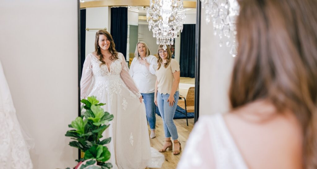 A bride in a lace wedding dress smiles at her reflection while two women react excitedly behind her in a bridal boutique.