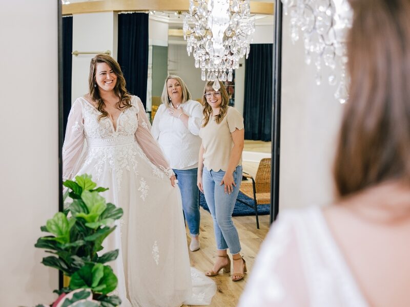 A bride in a lace wedding dress smiles at her reflection while two women react excitedly behind her in a bridal boutique.