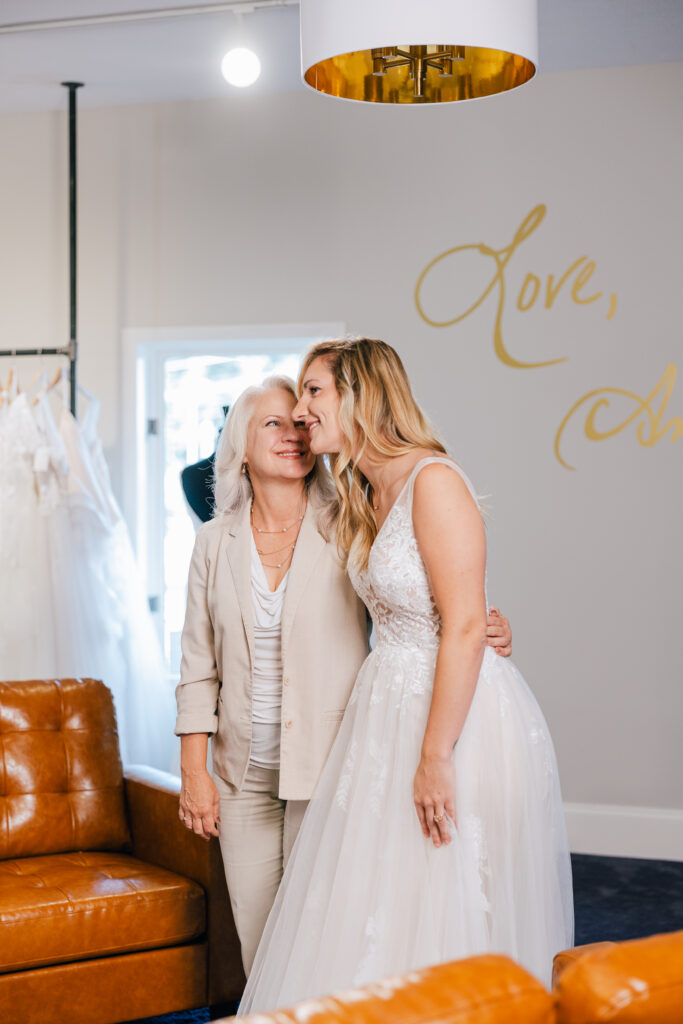 A bride in a lace and tulle wedding gown leans affectionately toward an older woman inside a bridal boutique.