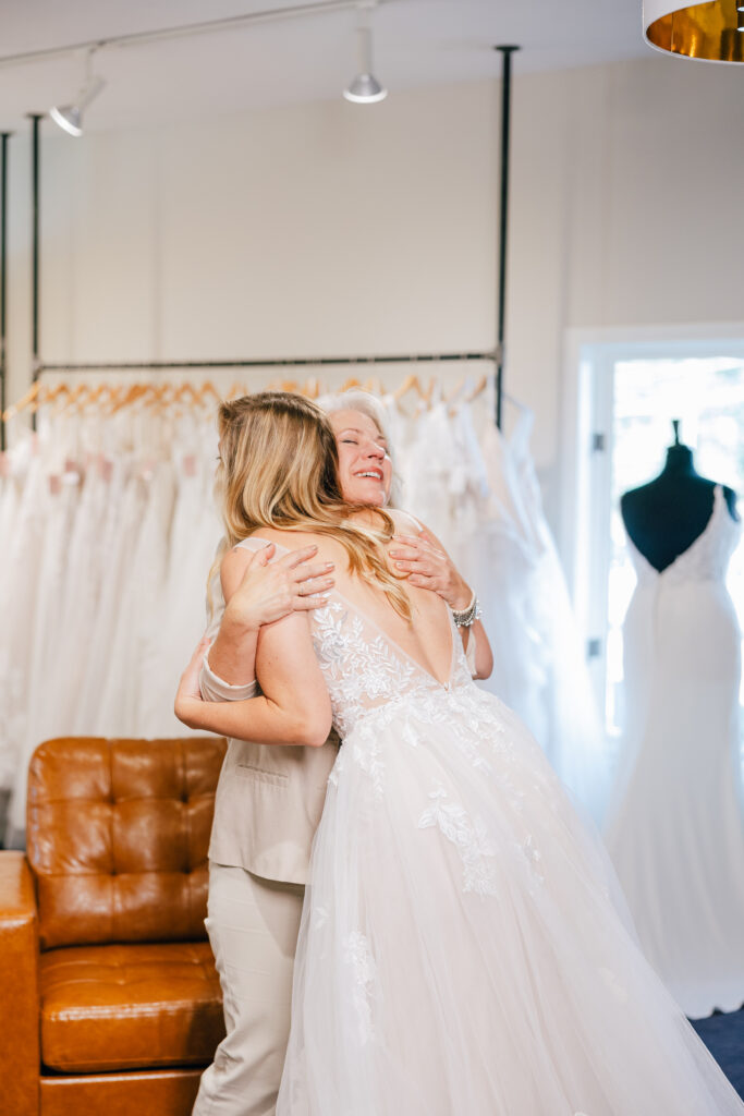 A bride in a lace wedding gown shares a joyful hug with an older woman beside racks of white dresses in a bridal shop.