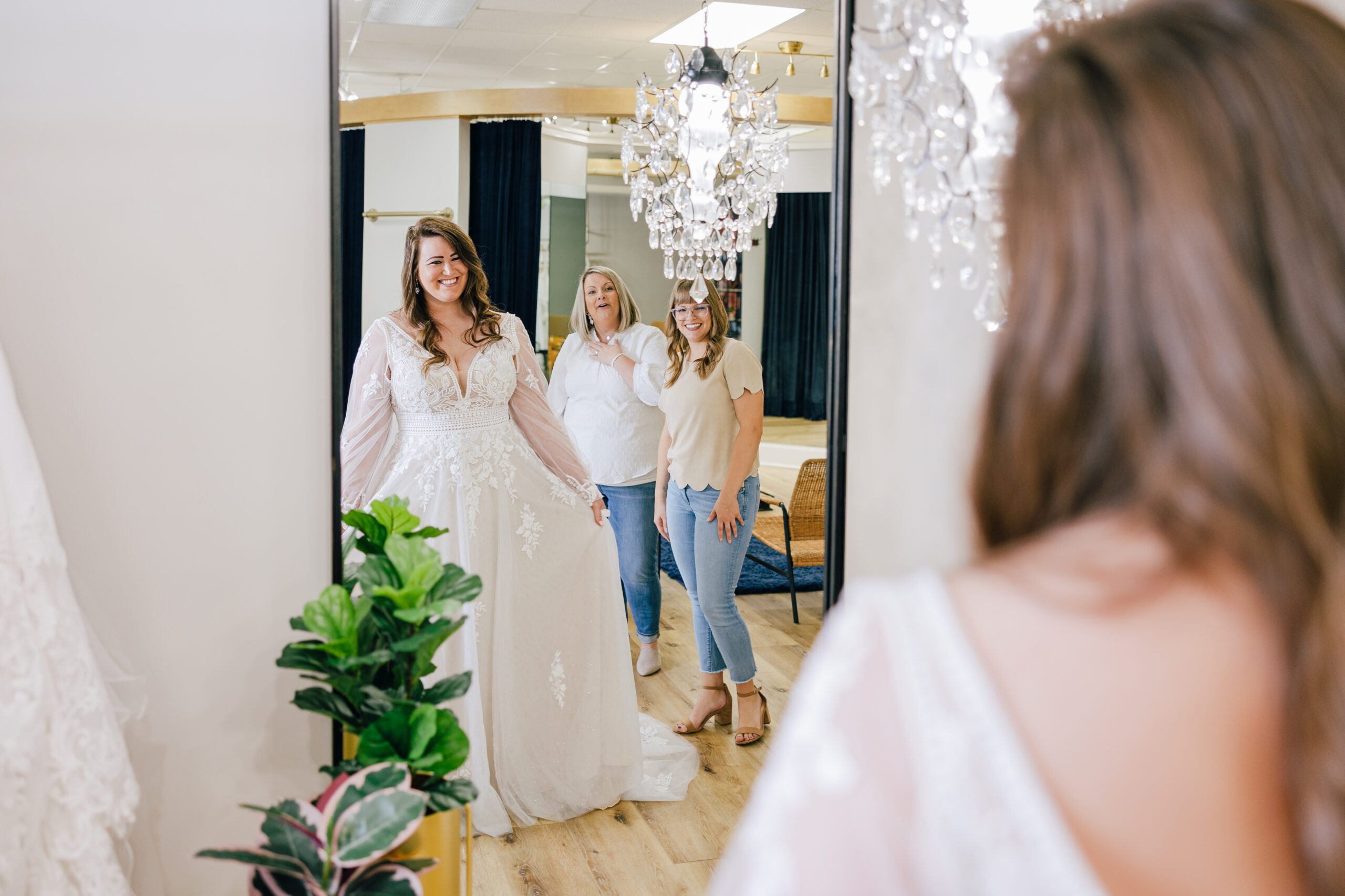 A smiling woman in a long-sleeved lace wedding gown looks into a large mirror while two women react happily behind her in a bridal fitting room.