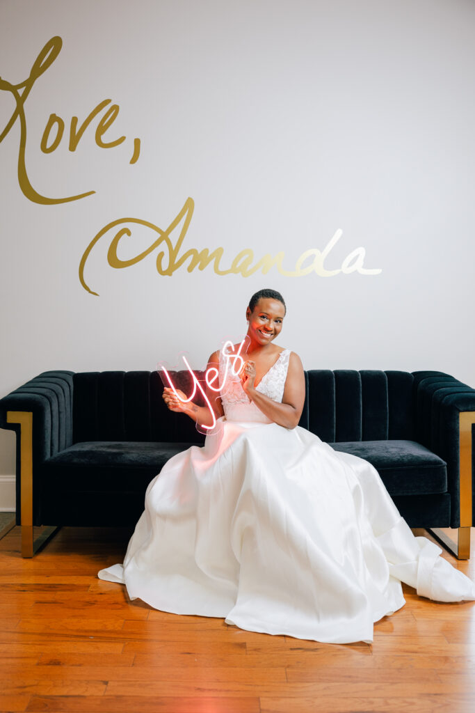 A bride in a white wedding gown sits on a dark velvet sofa while holding a glowing pink “Yes” sign.