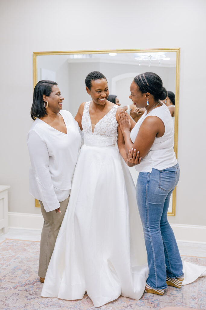 A bride in a sleeveless white wedding gown smiles and laughs with two women beside her in front of a large mirror.