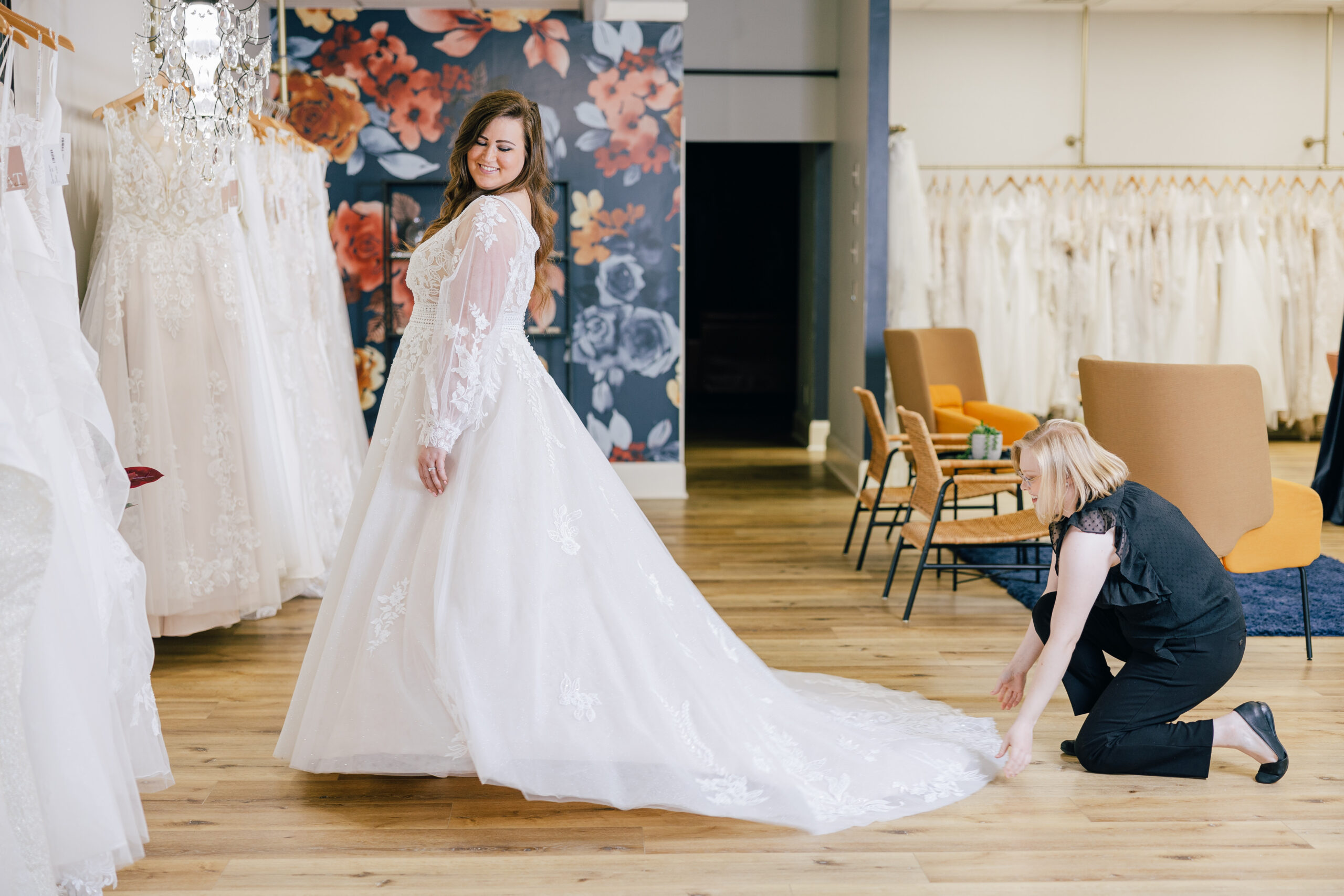 A smiling woman models a long-sleeved lace wedding gown while another woman kneels to arrange the train in a bridal boutique.