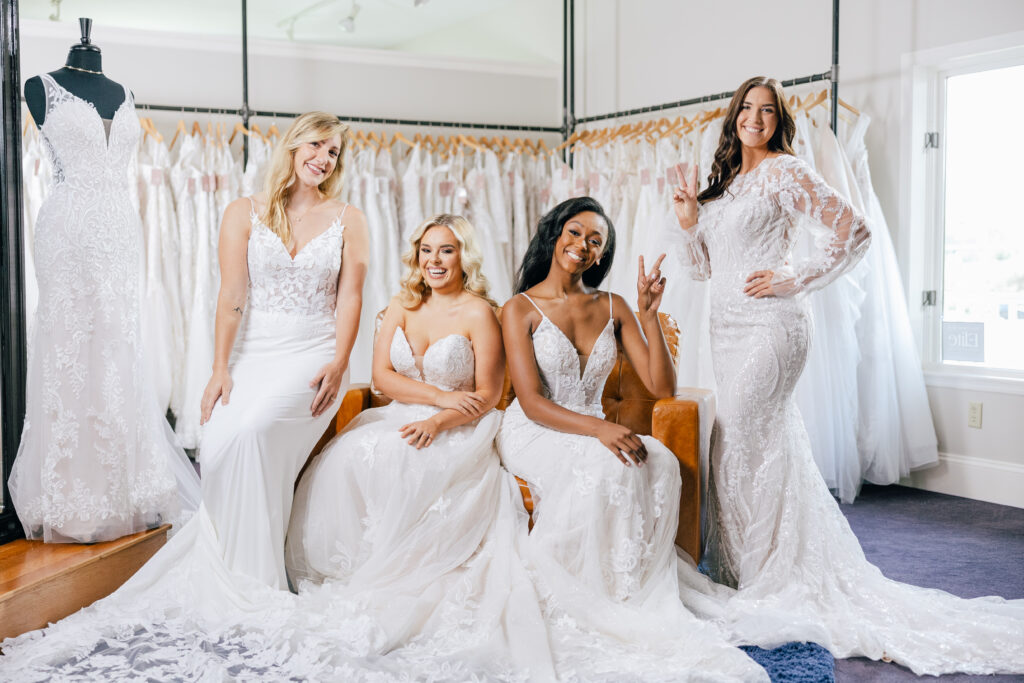 Four smiling women model different white lace wedding gowns together inside a bridal boutique.