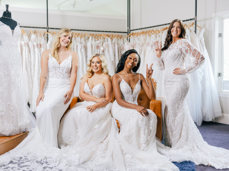 Four smiling women model different white lace wedding gowns together inside a bridal boutique.