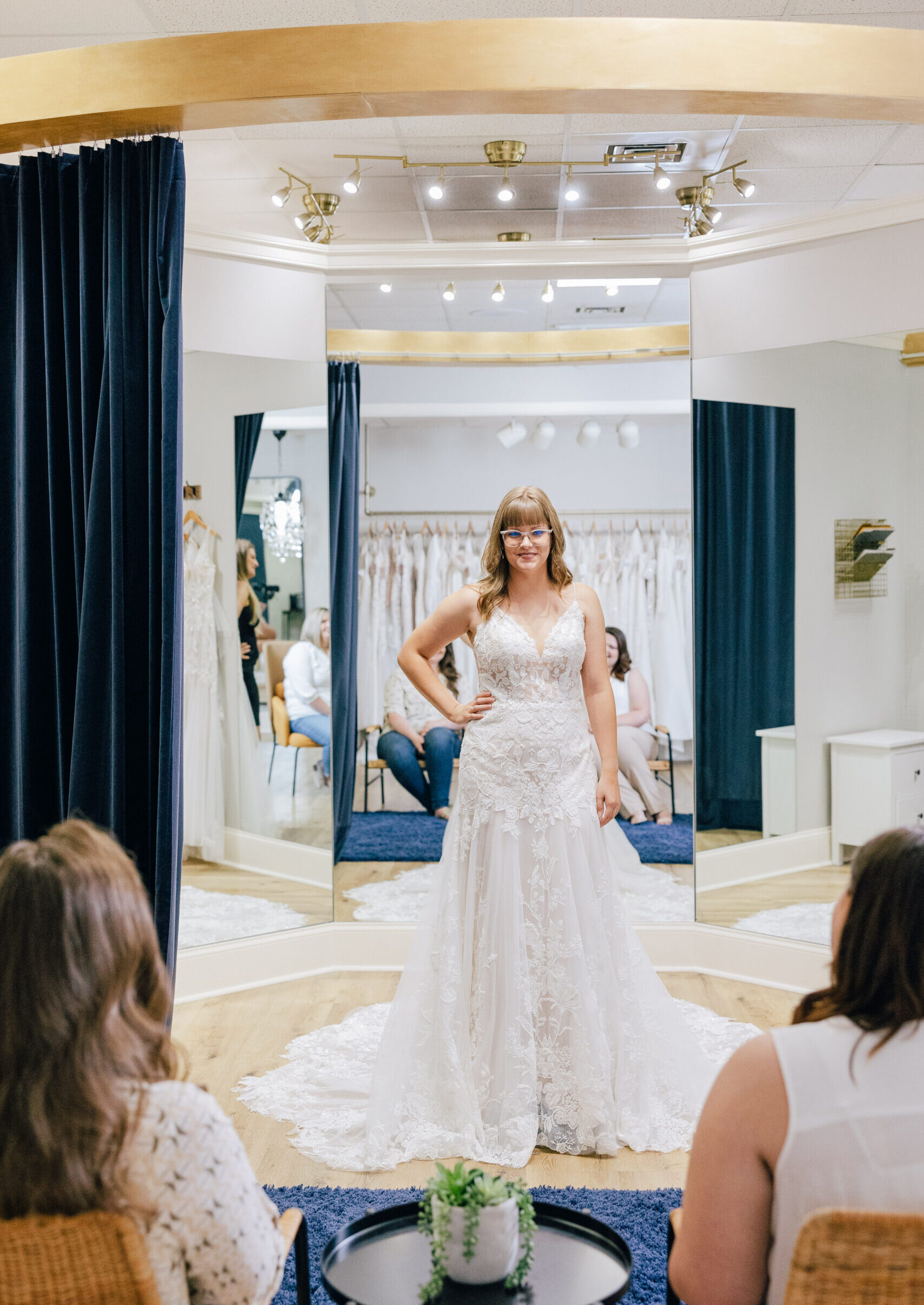 A woman wearing a lace wedding gown poses in front of large mirrors while two seated women watch from the fitting area.
