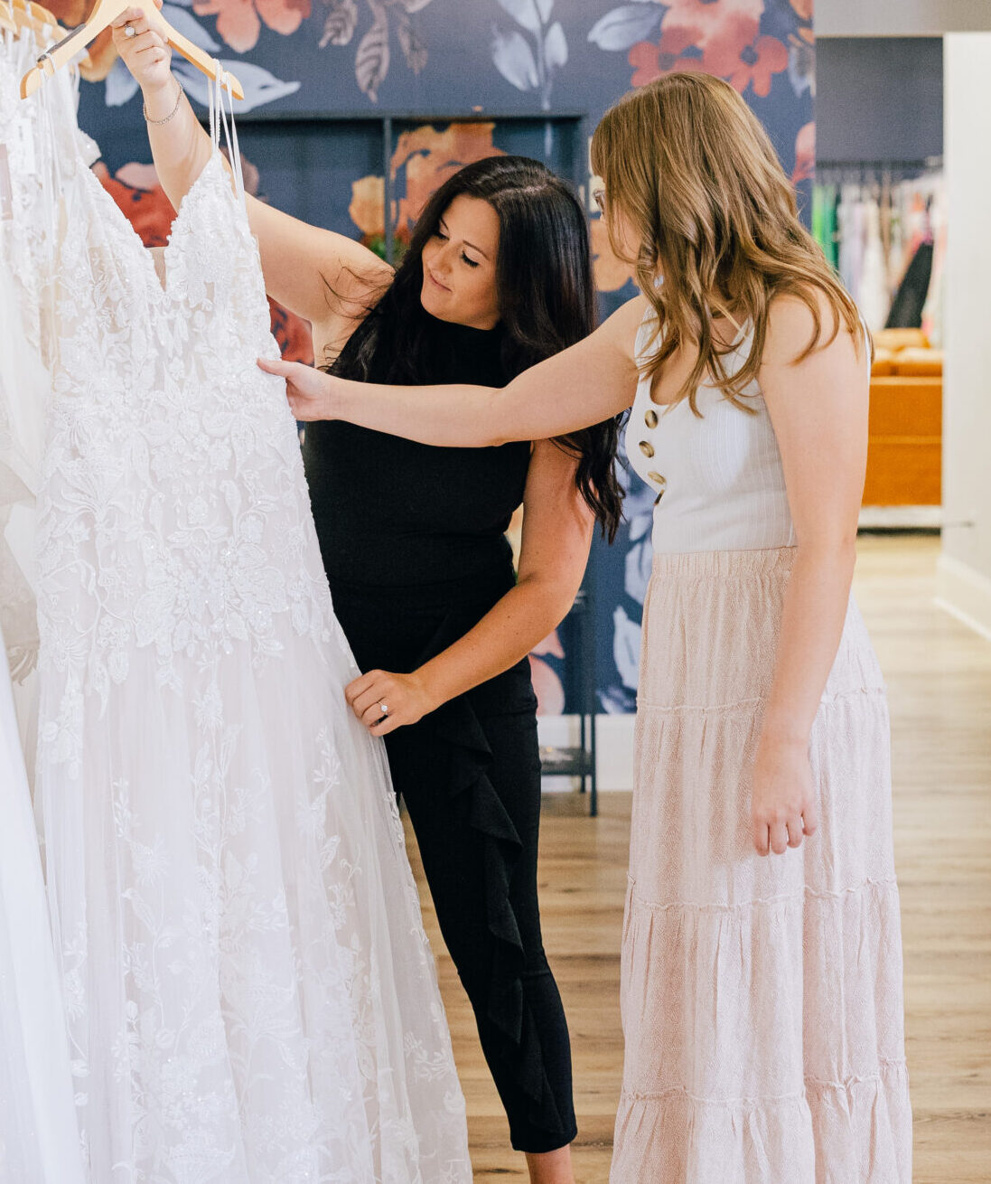 Two women examine the lace details of a white wedding dress hanging among other gowns in a bridal boutique.