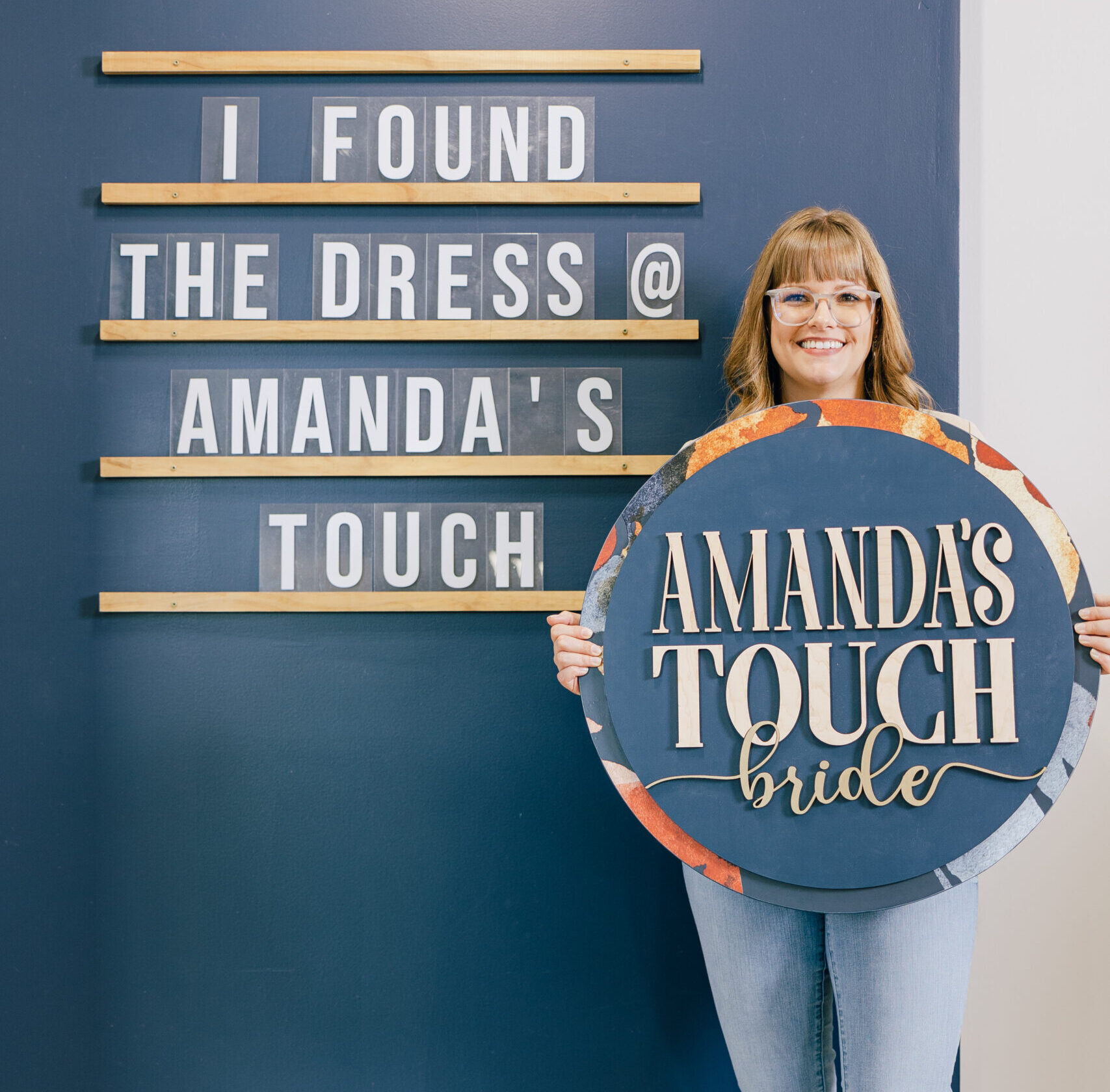 A smiling woman holds a round “Amanda’s Touch bride” sign beside a wall display reading “I found the dress @ Amanda’s Touch.”