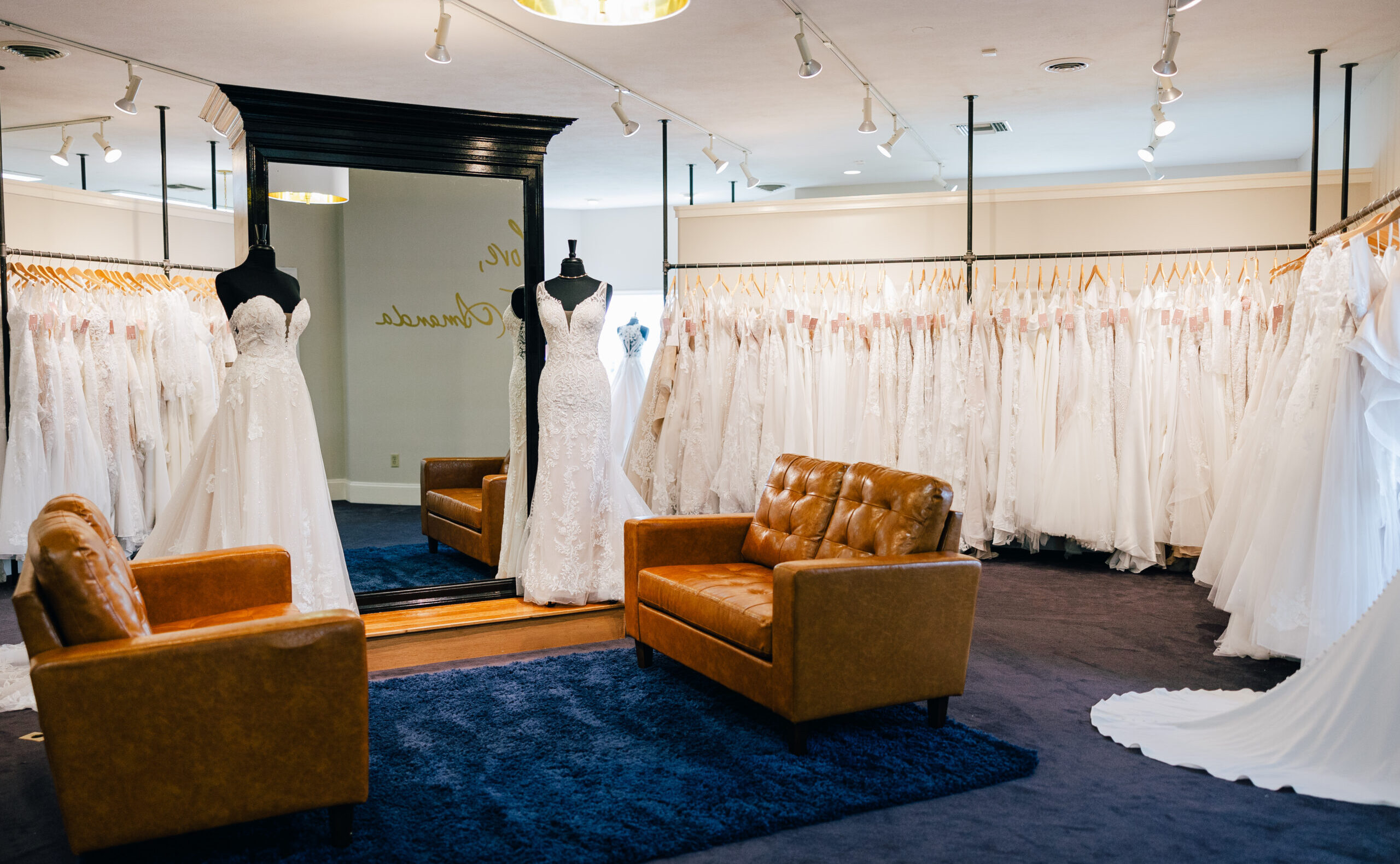 A bridal boutique showroom features rows of white wedding gowns, two dress mannequins, a large mirror, leather seating, and a blue area rug.