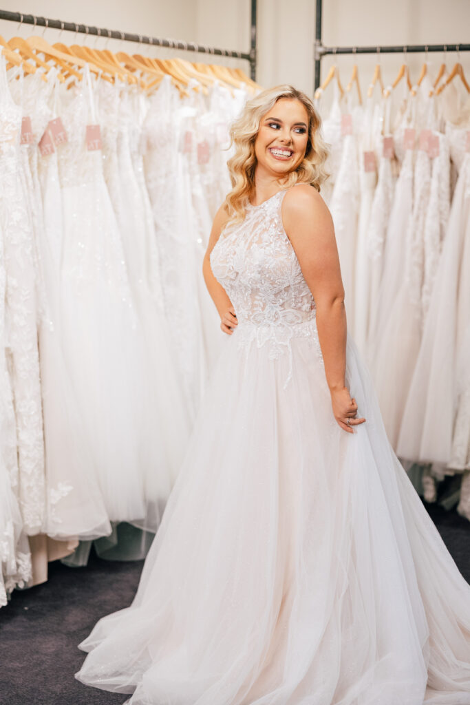 A smiling bride models a sleeveless lace wedding dress with a flowing tulle skirt in front of racks of bridal gowns.