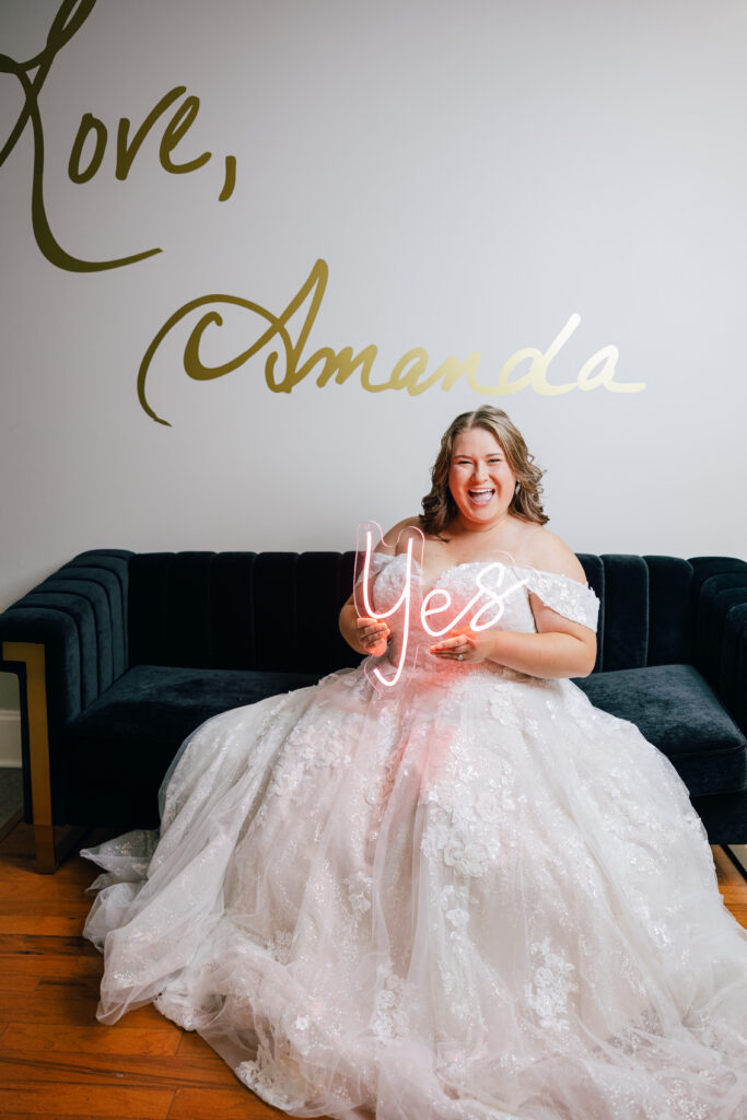 A bride in an off-the-shoulder lace ball gown sits on a dark velvet sofa while holding a glowing “Yes” sign.