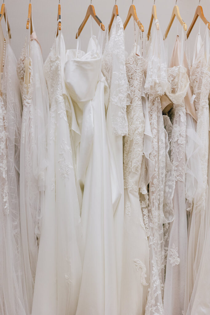 A collection of white and ivory wedding dresses with lace, beading, and satin details hangs on wooden hangers.