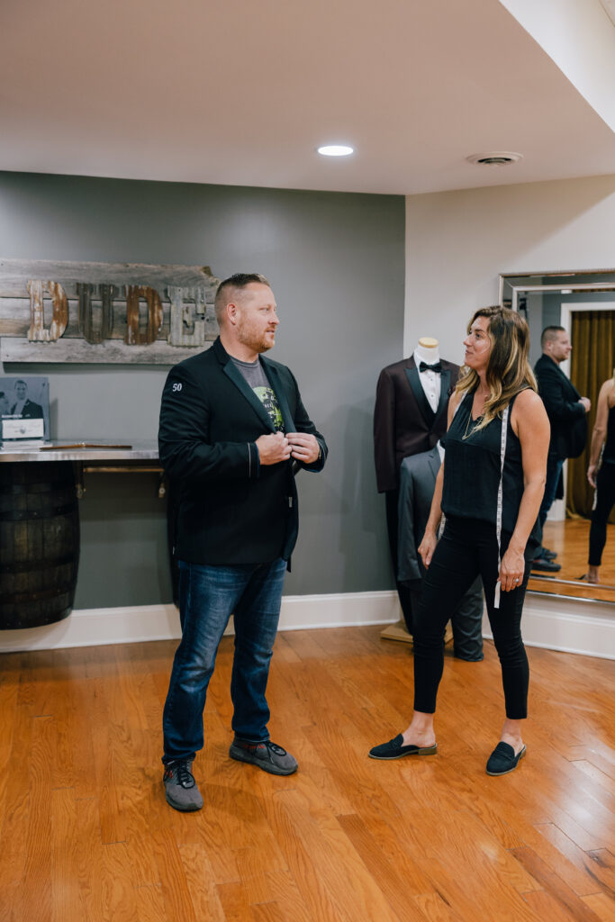 A man in a black blazer and jeans talks with a woman wearing a measuring tape in a formalwear shop beside a tuxedo mannequin.