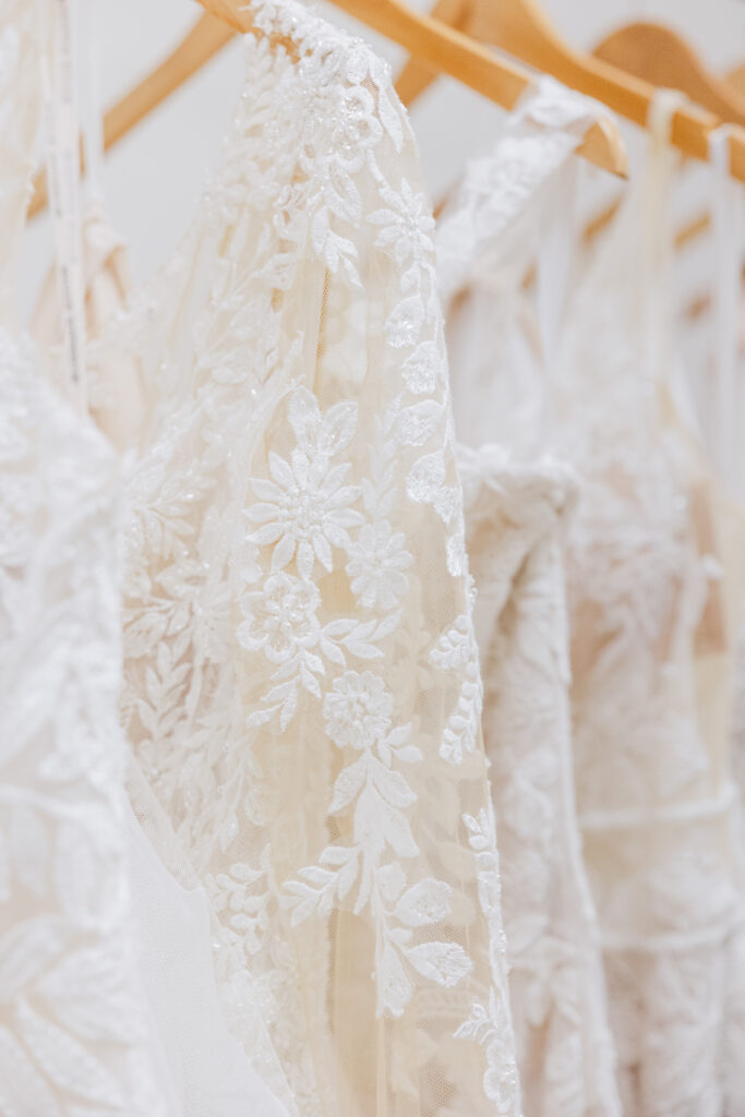 A close-up shows floral lace and beaded details on wedding dresses hanging together on a bridal rack.