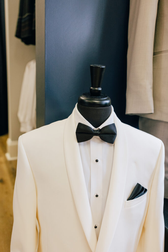 A white tuxedo jacket is displayed on a mannequin with a black bow tie, white dress shirt, and black pocket square.