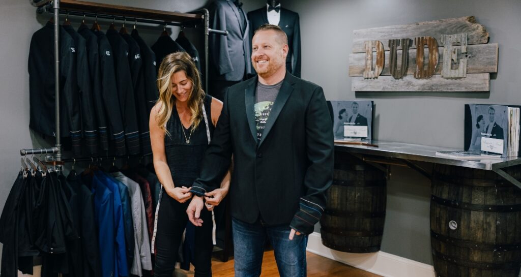 A woman with a measuring tape adjusts the sleeve of a man’s black blazer in a formalwear shop lined with suits.