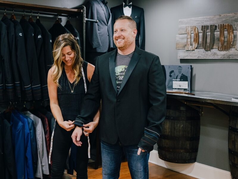 A woman with a measuring tape adjusts the sleeve of a man’s black blazer in a formalwear shop lined with suits.