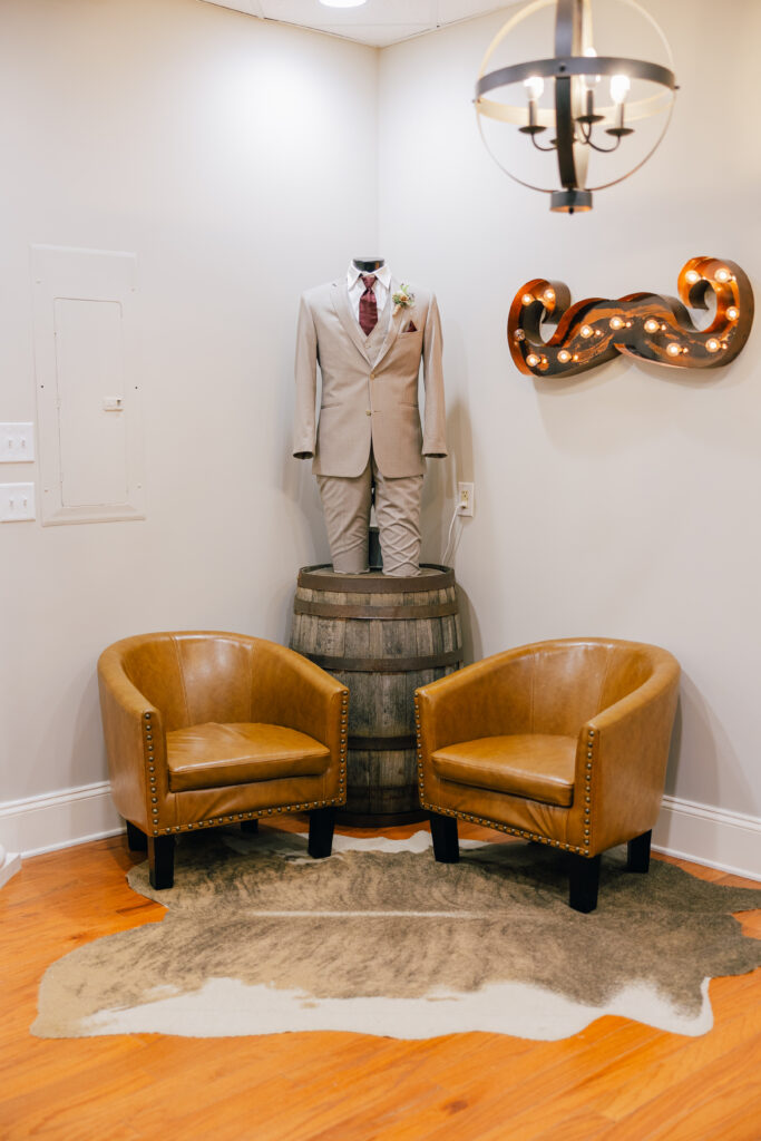A light beige three-piece suit with a burgundy tie and boutonniere is displayed on a mannequin above a wooden barrel between two leather chairs.