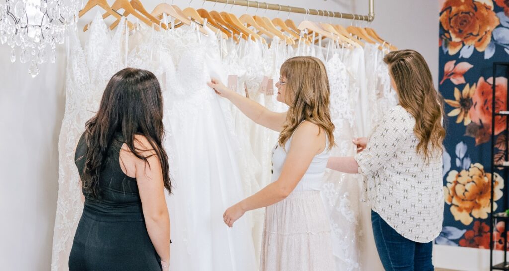 Three women browse a rack of white wedding dresses, examining different lace and fabric styles.
