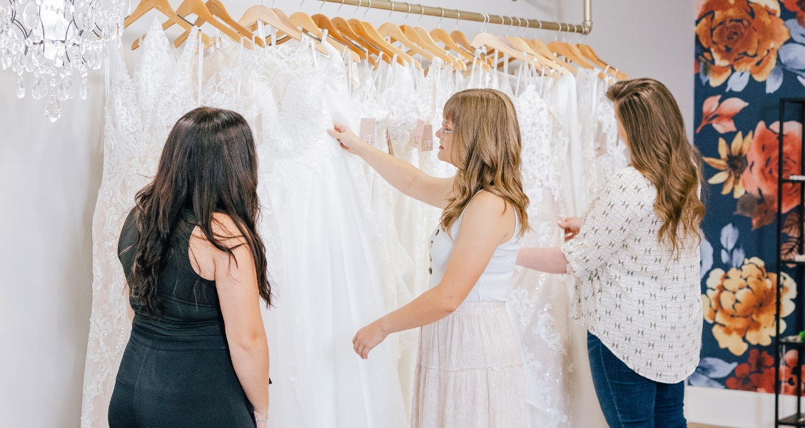 Three women browse a rack of white wedding dresses, examining different lace and fabric styles.