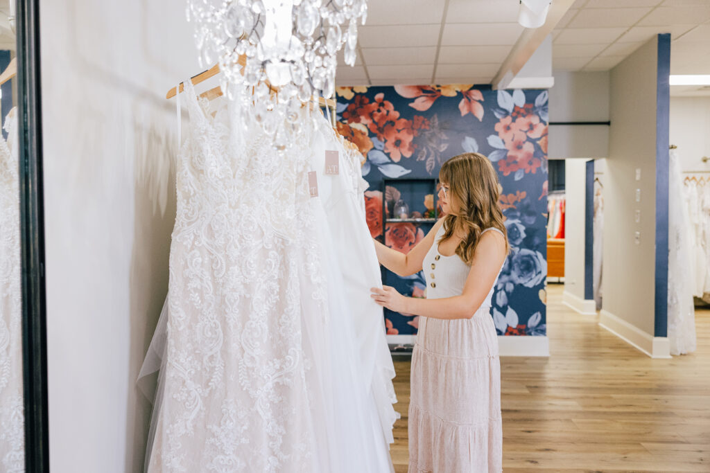 A woman looks through hanging wedding dresses in a bridal boutique with floral wallpaper and a crystal chandelier.