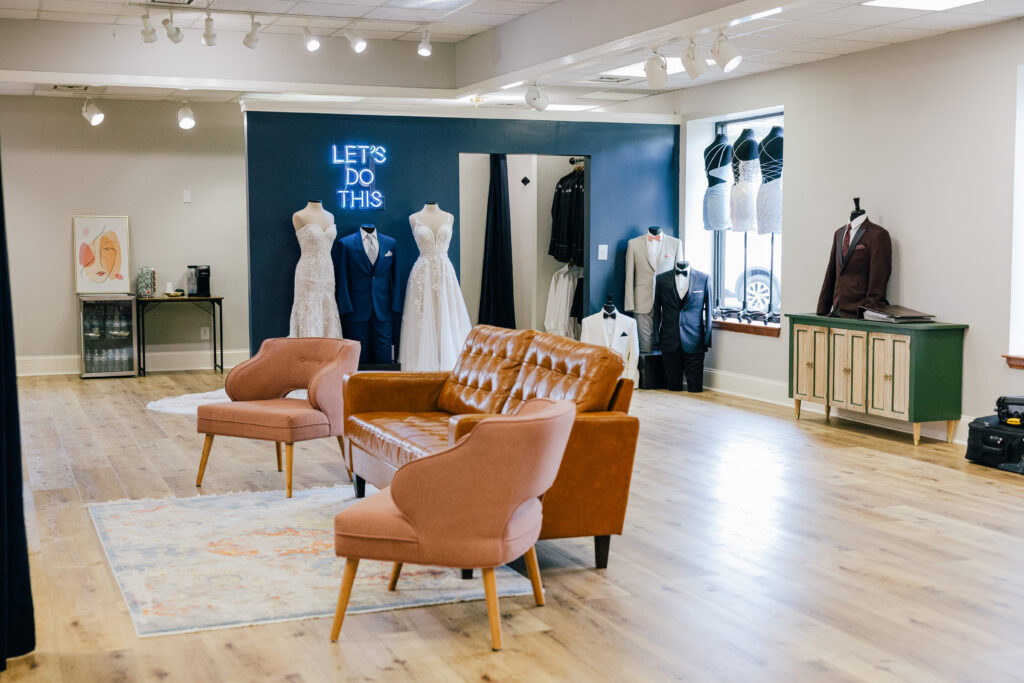 A spacious formalwear showroom features wedding dresses, suits, tuxedos, lounge seating, and a blue neon sign reading “LET’S DO THIS.”