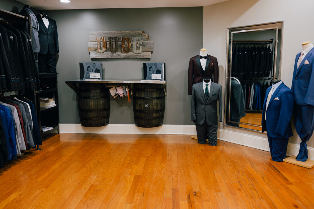 A formalwear showroom displays burgundy, gray, and blue suits on mannequins beside racks of jackets, a large mirror, and wooden barrel decor.